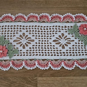 May include: A rectangular, handmade crochet doily with a white openwork center and a scalloped border in shades of pink and cream. The doily features floral accents with pink flowers and green leaves, perfect for home decor.
