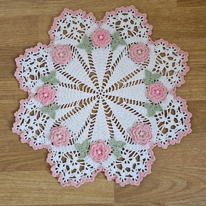 May include: White crocheted doily with a delicate floral design. The doily features pink flowers and green leaves around the edges.