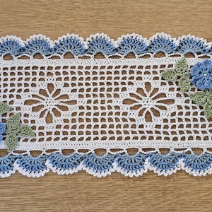 May include: A rectangular, handmade crochet doily. The center is white with a floral pattern. The border is blue and white, with scalloped edges. Blue flowers and green leaves are placed on the doily.
