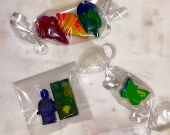 May include: Three small bags of crayons, each containing different shapes. The first bag contains a red heart, an orange ice cream cone, and a green popsicle. The second bag contains a blue Lego figure and a green Lego brick. The third bag contains a green butterfly.