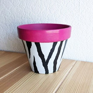 May include: A small terracotta planter painted with a black and white zebra stripe pattern. The top rim of the planter is painted a bright pink.