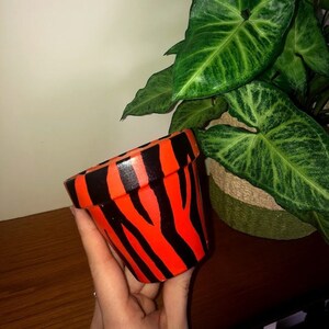 May include: A small terracotta pot painted with black and orange zebra stripes. The pot is being held in a hand.