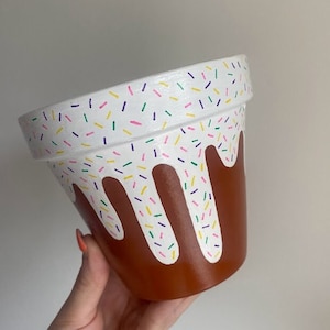 Op de afbeelding: Een terracotta bloempot geschilderd met witte glazuur en kleurrijke sprinkels. De glazuur druipt langs de zijkanten van de pot.