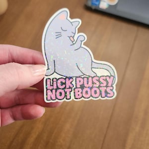 Puede incluir: Una pegatina con un gato morado claro lamiéndose la pata. El gato tiene un acabado brillante e iridiscente. Debajo del gato está el texto "LICK PUSSY NOT BOOTS" en letras rosas y blancas.