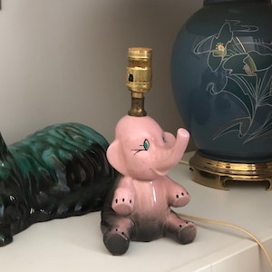 May include: A ceramic lamp base shaped like a pink elephant with black feet and ears, positioned next to a dark green and black ceramic cat figurine and a large blue table lamp with a pleated shade and floral gold-lined graphic details.