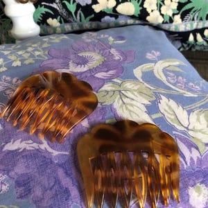 May include: Two hair combs crafted from brown and amber tortoiseshell-patterned material. Each comb features a scalloped top edge and a series of long, tapered teeth designed for securing hair, showcasing a smooth, glossy finish throughout.