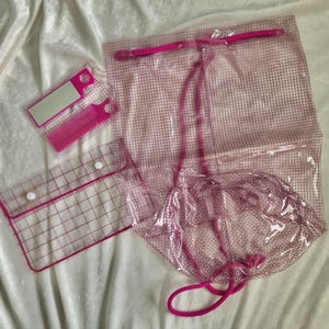 May include: A collection of clear, pink-trimmed, plastic accessories. Includes a drawstring bag, a small pouch with snaps, and two rectangular tags. The items are arranged on a white, textured surface.