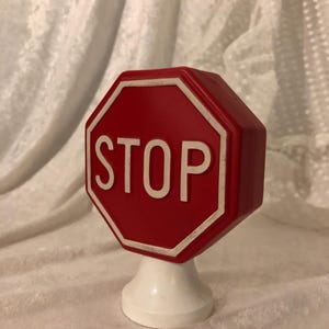 May include: A red, octagonal "STOP" sign with white lettering and a white border. The sign is mounted on a white, pedestal-style base. The sign is a decorative item.