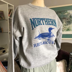 May include: A heather grey crewneck sweatshirt featuring a graphic print of a loon swimming on water. The text "NORTHERN" is printed in teal and dark blue above the loon, and the word "REFLECTION" is printed in dark blue below the loon.
