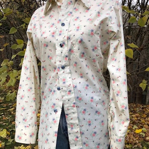 May include: Cream-coloured button-down shirt with a floral pattern in pink, blue, and green. The long-sleeved shirt has a classic collar and a single pocket. The shirt is unbuttoned, revealing blue jeans.