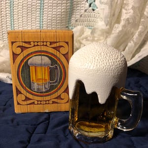 May include: A clear glass beer mug with a white foam top, beside a wooden box featuring a beer mug illustration. The mug contains a golden liquid. The box has a decorative border and a rainbow design.