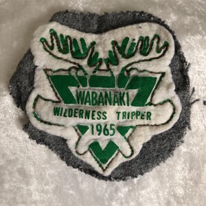 May include: A vintage patch with the text "WABANAKI WILDERNESS TRIPPER 1965" in green and white. The patch features a shield shape with moose antlers and a grey border. The patch is likely from a wilderness or outdoor adventure.