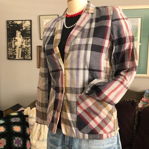 May include: A multi-coloured plaid blazer in navy, red, yellow, and white is displayed on a mannequin. The blazer features a classic collar, a single-button closure, and two front pockets. The mannequin also wears a black top, denim jeans, and a red and white beaded necklace.