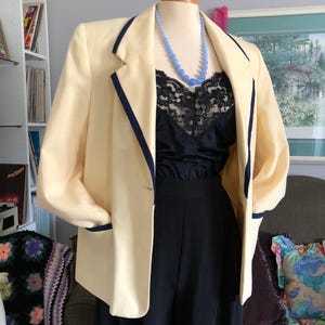 May include: A cream-colored blazer with navy blue trim and a black top and pants. A light blue beaded necklace is worn with the outfit. The blazer has a classic, tailored design.
