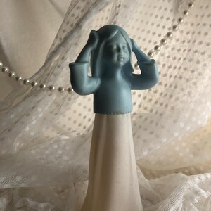 May include: A matte figurine of a person with long hair, featuring a light blue upper body and a long, off-white, flared skirt. The figure has both arms raised with hands touching the sides of the head, standing in a vertical, upright position.