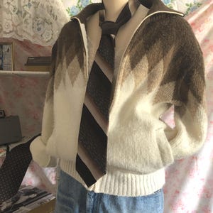 May include: A vintage white and brown zip-up sweater with a geometric pattern, paired with a striped tie. The sweater is displayed on a mannequin, showcasing a retro style. The tie features brown and beige stripes.
