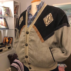 May include: A vintage bomber jacket in shades of gray, black, and tan. The jacket features a button-up front, pockets, and decorative patches on the shoulders. The jacket is displayed on a mannequin.