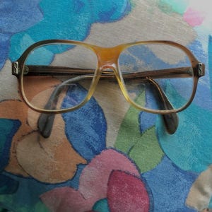 May include: Vintage spectacles with a light brown frame and dark brown arms. The lenses are clear. The glasses are resting on a floral patterned fabric with blue, pink, and orange hues.