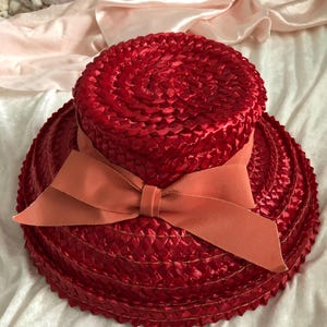 May include: A bright red sun hat with a wide brim and a matching orange bow. The hat's crown features a spiral pattern, and the bow is a muted orange. The hat is made of woven material, ideal for sun protection and style.