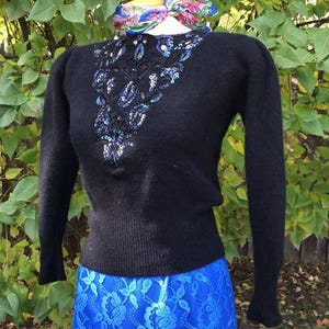 May include: A black sweater with a beaded neckline and long sleeves is worn with a bright blue lace skirt. A colourful scarf is tied around the neck. The outfit is displayed outdoors with a green leafy background.