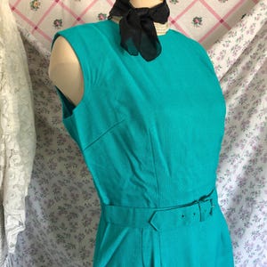 May include: A sleeveless, teal-colored dress with a matching belt and a black scarf tied around the neck. The dress is displayed on a mannequin. The background features floral patterned fabric.