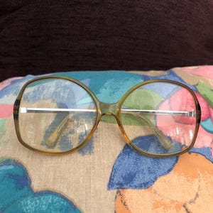 May include: Vintage spectacles with a light brown frame and clear lenses. The glasses have a rounded rectangular shape and silver-coloured metal arms. The frames are resting on a colourful floral patterned pillow.