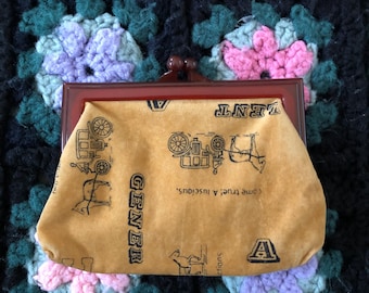 May include: A vintage brown purse with a rectangular frame and a clasp. The purse's body is mustard-colored and features black illustrations and text. The purse rests on a dark, multi-colored crocheted blanket.