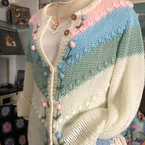 Peut inclure: Cardigan vintage tricoté à la main, crème, rose, bleu et vert. Le pull présente un motif rayé sur la poitrine et les manches, avec des broderies florales et des boutons en bois. Le cardigan est présenté sur un mannequin.