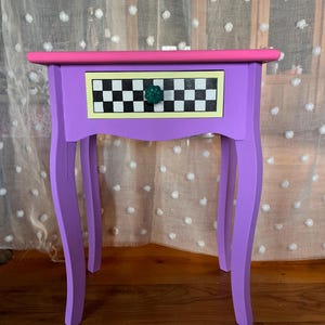 May include: A small side table with a bright pink top and a purple frame. The single drawer features a black and white checkered pattern bordered in yellow, accented by a round, dark green, textured knob. The table stands on four curved legs.
