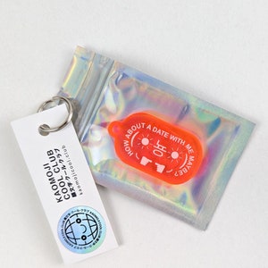 Könnte beinhalten: Ein roter Plastik-Schlüsselanhänger mit dem Text "HOW ABOUT A DATE WITH ME MAYBE?" und einem Daumen-hoch-Emoji mit dem Text "육" in Koreanisch. Der Schlüsselanhänger befindet sich in einer durchsichtigen Plastiktüte.