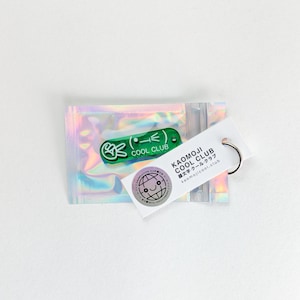 Könnte beinhalten: Ein grüner und weißer Schlüsselanhänger mit dem Text "COOL CLUB" und einem Peace-Zeichen-Emoji. Er ist an einem weißen Anhänger mit "KAOMOJI COOL CLUB" und einer Weltkugel-Grafik befestigt. Die Artikel befinden sich in einer holografischen Verpackung.