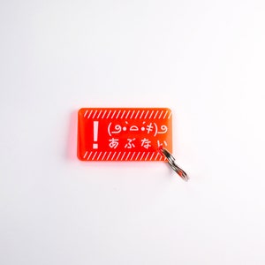 Könnte beinhalten: Ein leuchtend orangefarbener Schlüsselanhänger mit weißem Text, der "! (9.9. #)  あるな" lautet, und einem silbernen Metallring.