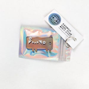 Könnte beinhalten: Ein brauner und weißer Charakter-Schlüsselanhänger mit Blumenmuster. Der Schlüsselanhänger befindet sich in einer durchsichtigen Plastiktüte mit holografischem, irisierendem Finish. Die Tüte ist an einem weißen Etikett mit dem Text "KAOMOJI COOL CLUB" und der Website "kaomojicool.club" befestigt.