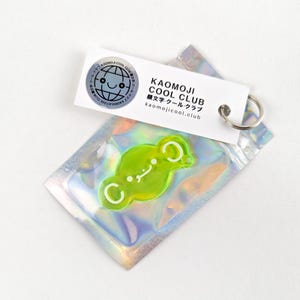 Könnte beinhalten: Ein grün-weißer Schlüsselanhänger mit einem Comic-Gesicht und dem Text "KAOMOJI COOL CLUB 顔文字・クール・クラブ kaomojicool.club".
