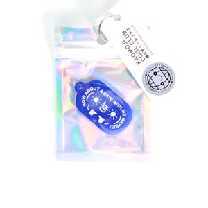 Könnte beinhalten: Ein blauer Schlüsselanhänger mit dem Text "HOW ABOUT A DATE WITH ME MAYBE?" befindet sich in einer holografischen Tasche. Der Schlüsselanhänger hat eine Sonne und ein Gesichts-Grafik. Ein weißes Etikett mit japanischem Text und einem Logo ist angebracht.