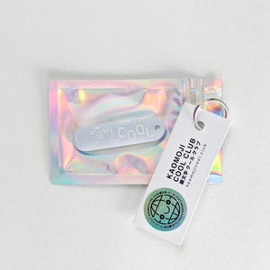 Könnte beinhalten: Ein hellblaues Schlüsselanhänger mit dem Wort "COOL" und einem lächelnden Gesichtsdesign, verpackt in einem holografischen, irisierenden Beutel. Ein weißes Etikett mit "KAOMOJI COOL CLUB" Text und Logo ist angebracht.