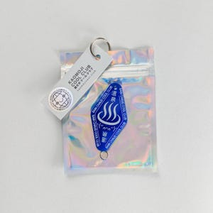 Könnte beinhalten: Ein blauer Schlüsselanhänger mit dem Text "BAIN CHAUD COOL CLUB" und einer Grafik von drei Dampflinien. Der Schlüsselanhänger ist an einem silbernen Ring und einem Anhänger mit dem Text "KAOMOJI COOL CLUB" befestigt. Die Artikel befinden sich in einer holografischen Tasche.