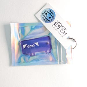 Könnte beinhalten: Ein lila, durchscheinender Schlüsselanhänger in einer holografischen Tüte mit einem Anhänger. Der Schlüsselanhänger hat ein einzigartiges Design mit weißen geometrischen Formen und dem Text "O&O". Auf dem Anhänger steht "KAOMOJI COOL CLUB" und "kaomojicool.club".