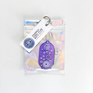 Könnte beinhalten: Ein lila Schlüsselanhänger mit weißen Blumen- und Sterndesigns, verpackt in einer transparenten, irisierenden Tasche. Der Schlüsselanhänger hat japanische Schriftzeichen und einen Metallring. Ein weißes Etikett mit dem Text "KAOMOJI COOL CLUB" ist angebracht.