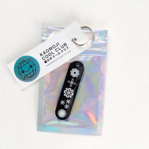 Könnte beinhalten: Ein schwarzer Schlüsselanhänger mit weißen Blumenmustern und dem Text "KAOMOJI COOL CLUB" auf einem weißen Anhänger. Der Schlüsselanhänger befindet sich in einer holografischen, irisierenden Tasche. Auf dem Anhänger steht auch die Website kaomojicool.club.