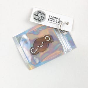 Könnte beinhalten: Ein brauner und weißer Schlüsselanhänger mit dem Text "C///W" und einem kleinen Kreis mit zwei Punkten. Der Schlüsselanhänger befindet sich in einer durchsichtigen Plastiktüte mit einem silbernen Reißverschluss. Die Tüte liegt auf einer weißen Oberfläche. Der Schlüsselanhänger ist auch an einem silbernen Schlüsselring mit einem Anhänger befestigt, auf dem "KAOMOJI COOL CLUB" in schwarzer Schrift und "顔文字クールクラブ" in japanischer Schrift steht. Der Anhänger hat auch einen kleinen Globus mit einem Smiley-Gesicht.