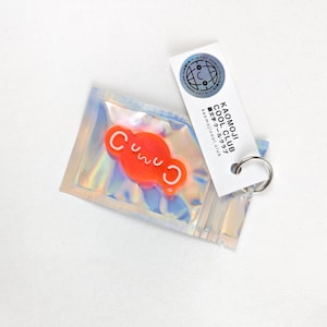 Könnte beinhalten: Ein kleiner orangefarbener Schlüsselanhänger mit dem Wort "Cuuu" in weißen Buchstaben. Der Schlüsselanhänger ist in einer durchsichtigen Plastiktüte mit silbernem Holographie-Finish verpackt. Die Tüte enthält auch ein weißes Etikett mit dem Text "KAOMOJI COOL CLUB" in schwarzen Buchstaben und dem Text "カオモジクールクラブ" in japanischer Sprache.