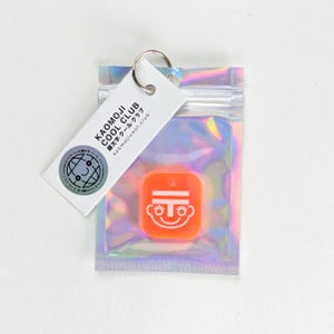Könnte beinhalten: Ein orangefarbener Schlüsselanhänger mit einer weißen Gesichtsgrafik in einem transparenten, schillernden Beutel. Der Schlüsselanhänger hat einen silbernen Ring und ein weißes Etikett mit dem Text "KAOMOJI COOL CLUB" und "kaomojicool.club". Ein kleiner Aufkleber mit einem Globus-Motiv ist ebenfalls angebracht.