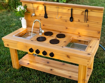 May include: A wooden outdoor play kitchen with a sink, stove, and storage shelf. The kitchen has a water faucet, black burner tops, and hanging utensils. A white vase with flowers sits on the counter. The play kitchen is on a green lawn.