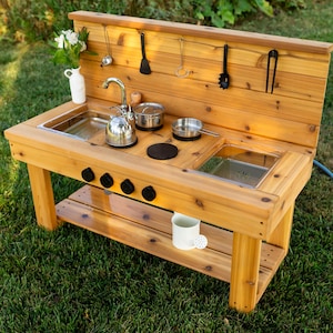 May include: A wooden outdoor play kitchen featuring a sink, a faucet, two metal basins, and four black knobs. The unit includes a backsplash with hanging hooks for kitchen utensils, a lower shelf with a white container, and metal cookware.