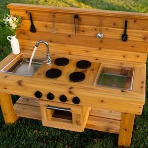 May include: A wooden outdoor play kitchen featuring a sink with a functional tap, four black circular stovetop burners, and a central oven compartment. The backsplash includes five hanging black kitchen utensils and a white vase with flowers.