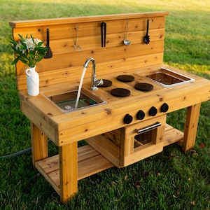 May include: A wooden outdoor play kitchen featuring a sink with a functional faucet, three black stovetop burners, and a lower oven compartment. The backsplash includes hooks holding black kitchen utensils, and a white vase with flowers sits nearby.
