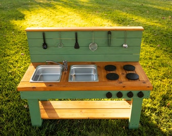 May include: A wooden outdoor play kitchen with a green painted backsplash and natural wood countertop. It features two stainless steel sinks, a faucet, and four black stovetop burners. Utensils hang above. The kitchen is set on a grassy lawn.