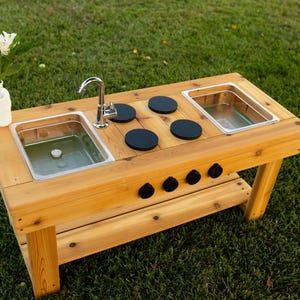 May include: A wooden outdoor play kitchen featuring a natural cedar finish, two stainless steel basins, a silver-toned metal tap, four black circular stovetop burners, and four black control knobs on the front panel, with a lower storage shelf.
