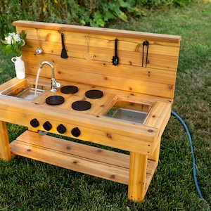 May include: A wooden outdoor play kitchen with a sink, stove, and storage shelf. The kitchen has a water faucet, black burner tops, and hanging utensils. A white vase with flowers sits on the counter. The play kitchen is on a green lawn.